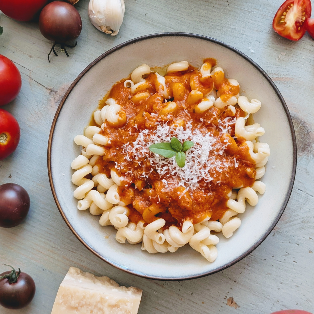Bowl of curly pasta topped with tomato sauce, grated Parmesan and a basil sprig, with tomatoes and garlic nearby.