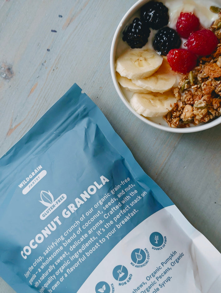 Top-down angled view of gluten-free Coconut Granola pouch labeled 'Coconut Granola' beside a bowl with banana and berries