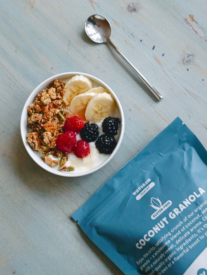 Top-down view of 9oz gluten-free coconut granola bag showcasing crunchy granola clusters beside a bowl topped with banana slices and berries on light blue wood