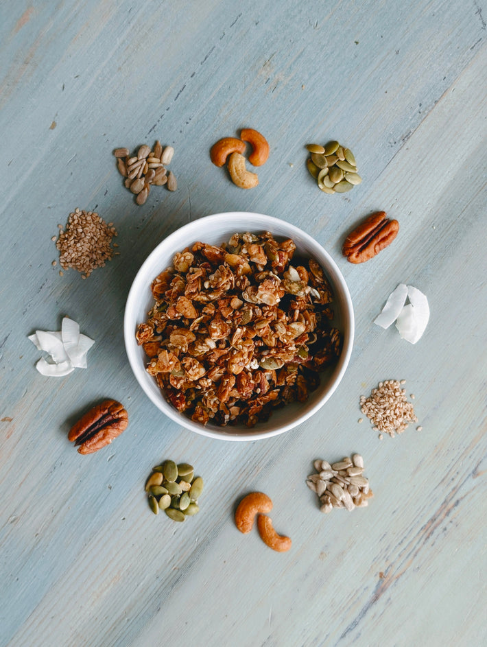 Overhead view of Gluten‑Free Coconut Granola (9oz) in a white bowl, showing toasted coconut, nut and seed clusters on pale blue wood