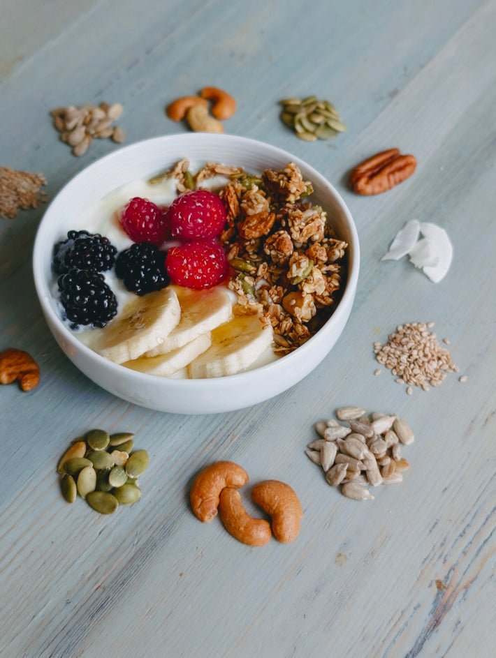 Top-down view of gluten-free coconut granola in a bowl with banana slices, raspberries, blackberries and seeds on light wood