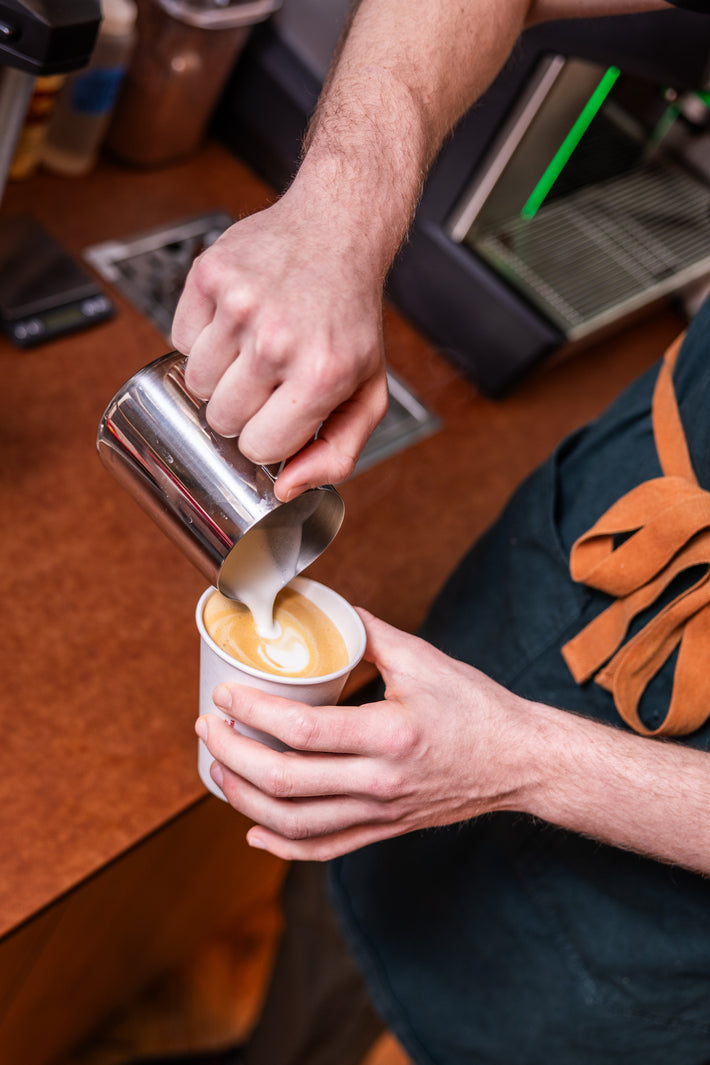 Barista pouring milk into a coffee cup on a wooden counter.