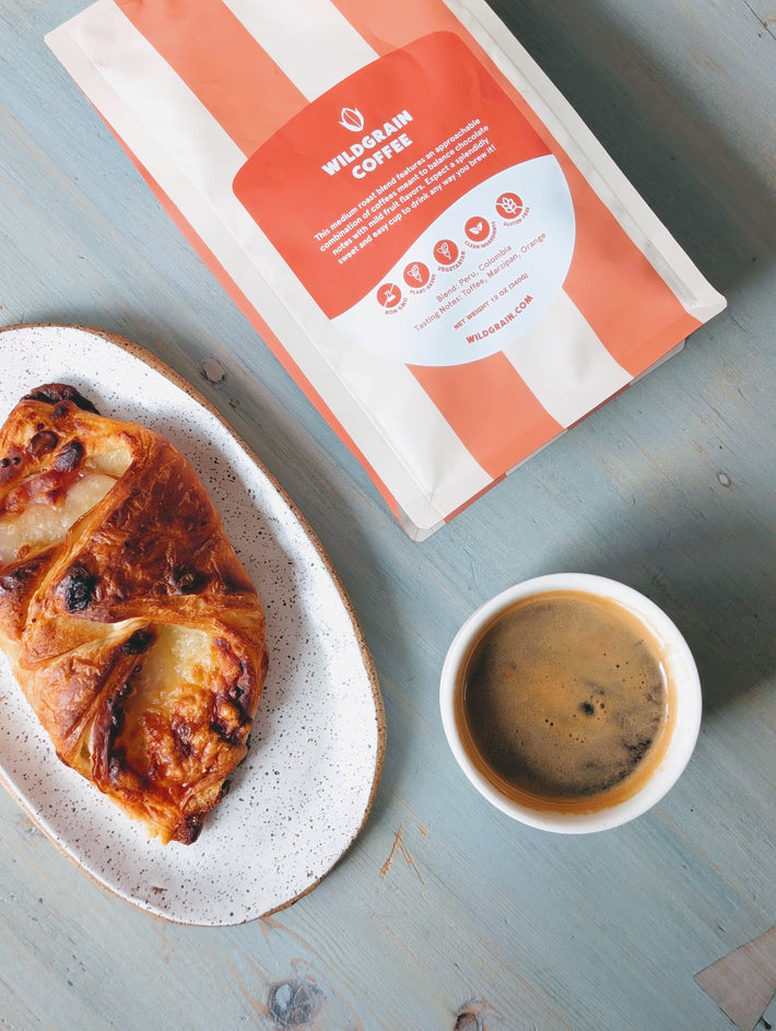 Top-down view of Wildgrain Coffee whole bean bag with coral-and-white packaging beside espresso and pastry on pale blue table
