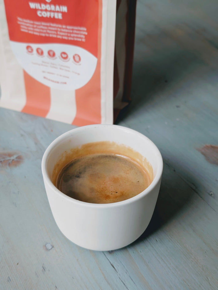 White ceramic cup of brewed coffee on a painted blue wood table with a Wildgrain coffee bag blurred in the background