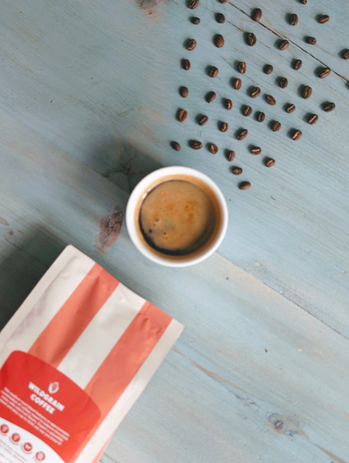 Top-down view of Wildgrain medium roast whole bean coffee bag with a cup of espresso and scattered beans on a light blue wooden table