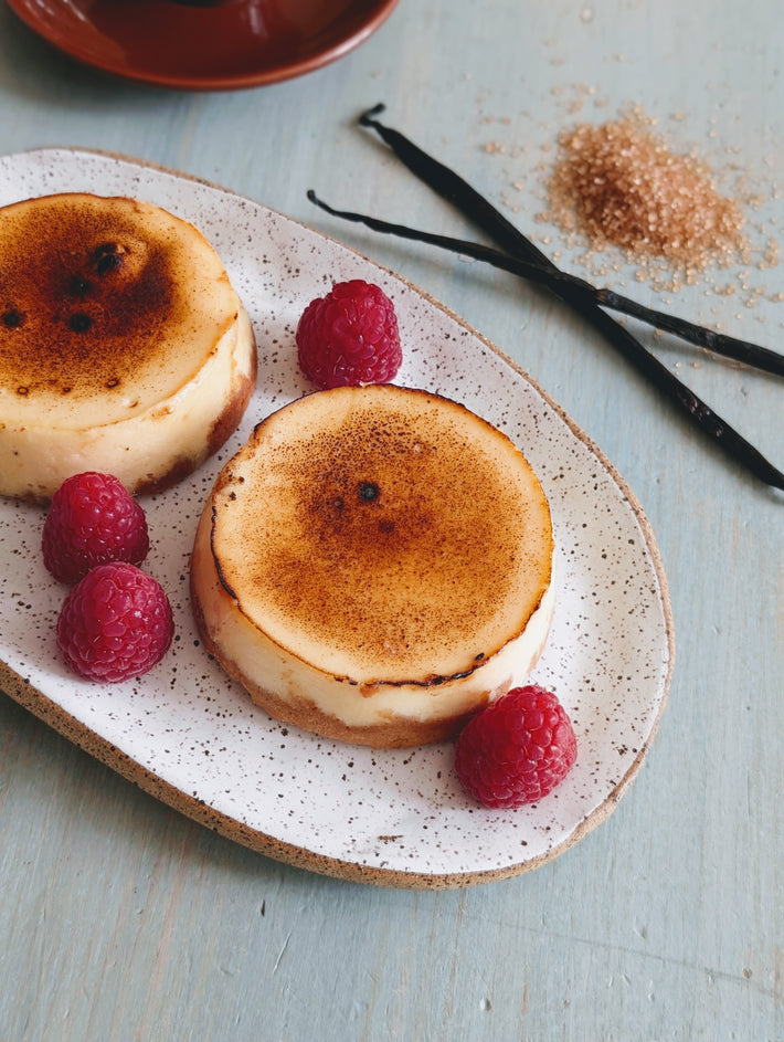 Two crème brûlée cheescakes on a plate with raspberries and vanilla beans on a light blue background