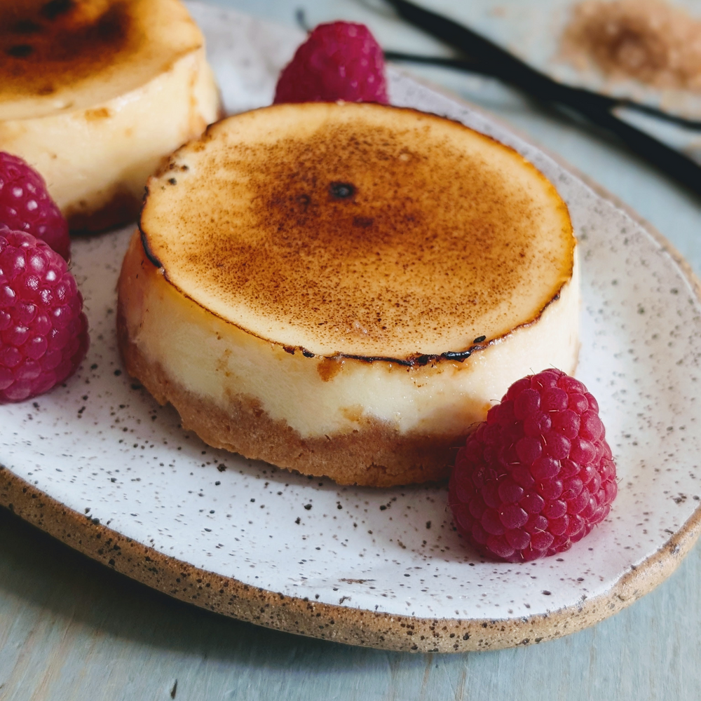 Two mini brûléed cheesecakes on a speckled plate, garnished with fresh raspberries