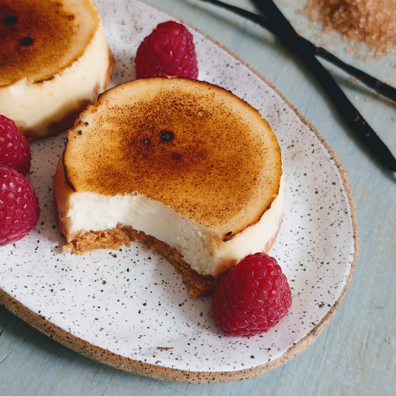 Mini bruleed cheesecake with a bite taken, on a speckled plate with raspberries and vanilla pods