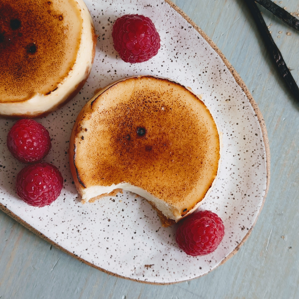Two mini brûlée cheesecakes on a speckled oval plate with raspberries; one cheesecake has a bite taken out.