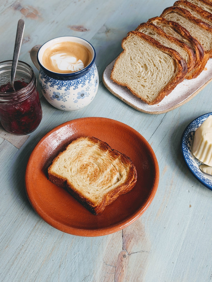A slice of toasted bread on a terracotta plate with a cup of coffee, a jar of jam, and a butter dish. Jam jar has text on it.
