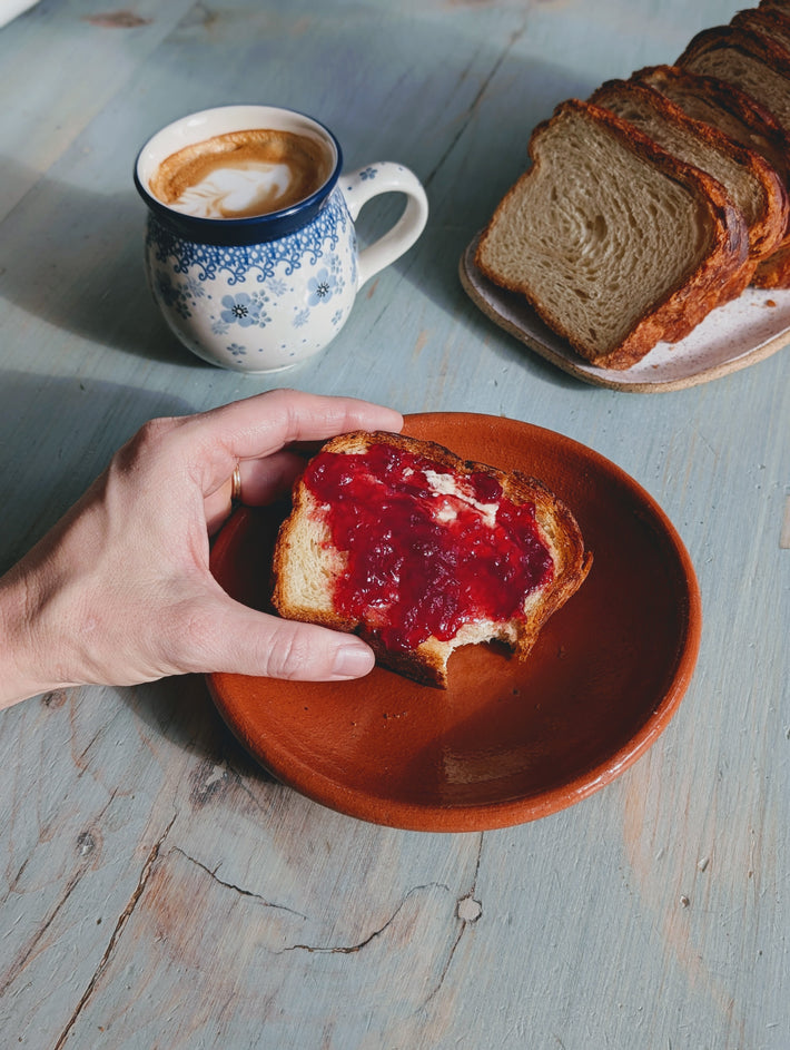 Hand holding a bitten slice of toast spread with red jam on an earthenware plate; sliced loaf and latte in a blue floral mug