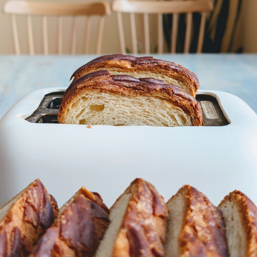 Two slices of braided challah bread warming in a white toaster, with sliced loaf pieces blurred in the foreground