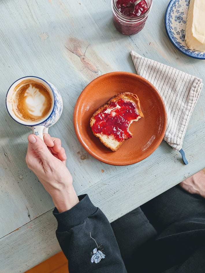 A hand holding a coffee cup next to a plate of toast with jam and a striped napkin.