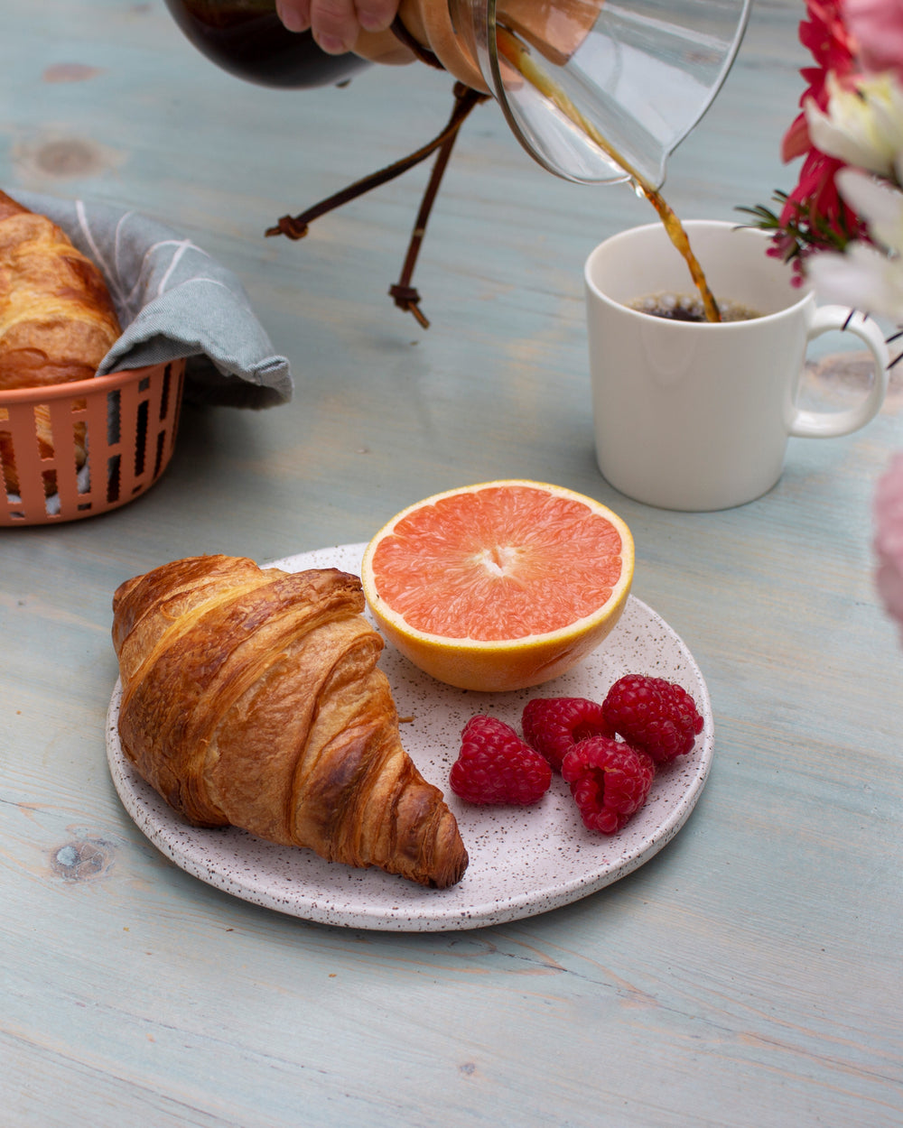 A croissant on a plate with raspberries and a grapefruit half, with coffee being poured in the background.