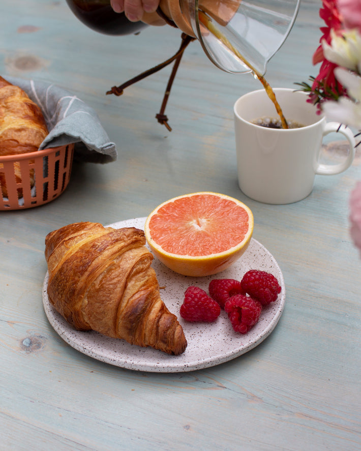 A croissant on a plate with raspberries and a grapefruit half, with coffee being poured in the background.