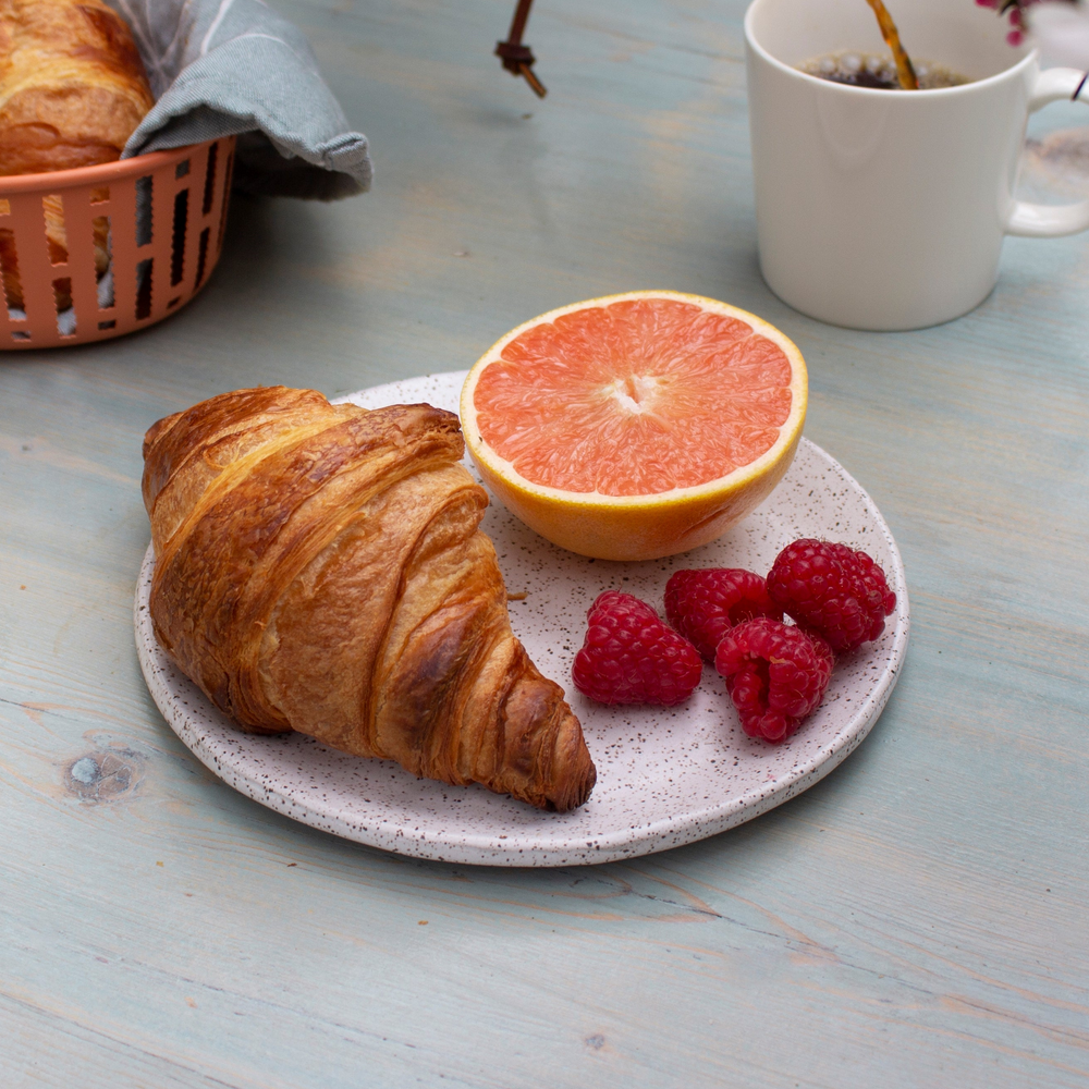 Croissant, halved grapefruit and three raspberries on a speckled plate beside a white coffee mug