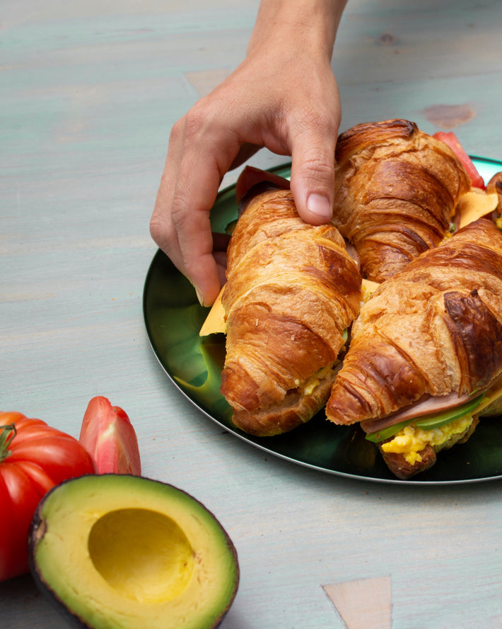 A hand reaching for croissants on a plate with avocado and tomatoes