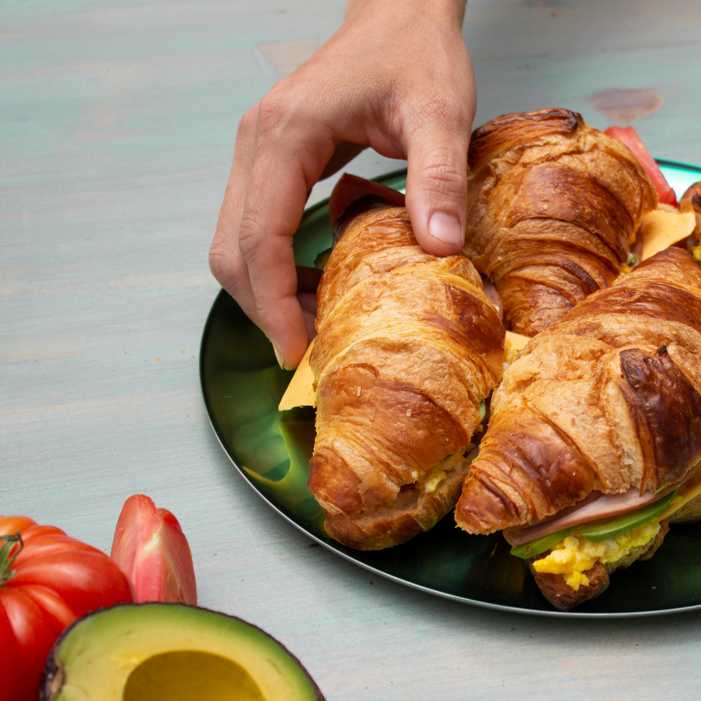 Hand picking a croissant sandwich with cheese and scrambled eggs on a green plate; avocado and tomato halves nearby.