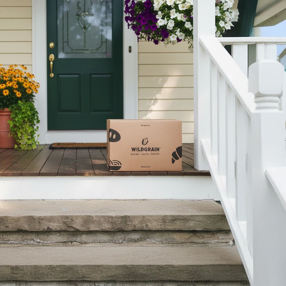 Cardboard Wildgrain delivery box on a front porch step, showing WILDGRAIN BREAD PASTA PASTRY