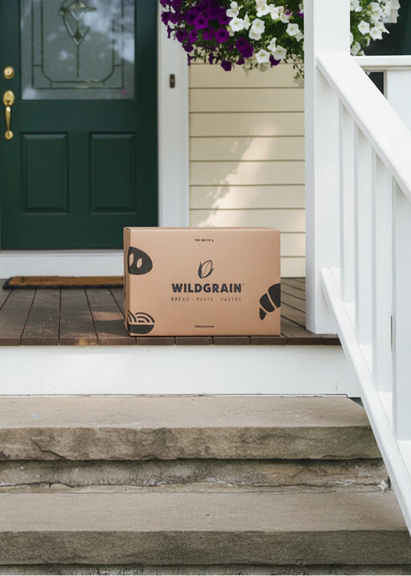 Cardboard Wildgrain delivery box on a front porch step, showing text WILDGRAIN BREAD · PASTA · PASTRY wildgrain.com