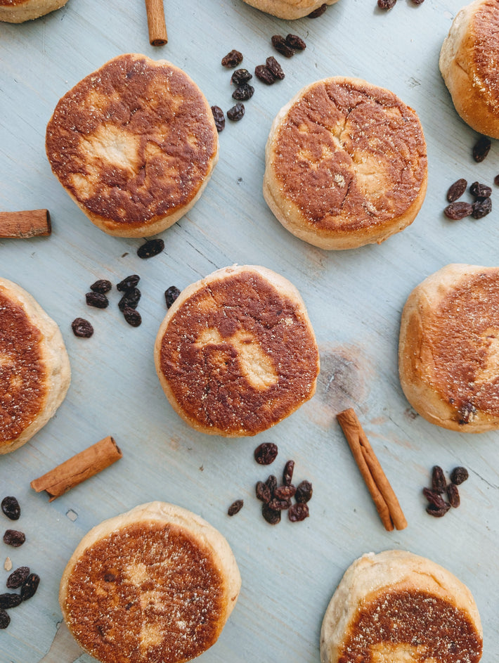 Toasted English muffins arranged on a pale blue wooden board, with scattered raisins and cinnamon sticks.