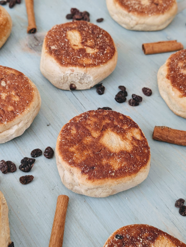 Toasted crumpets with browned tops on a pale blue board, scattered cinnamon sticks and raisins.