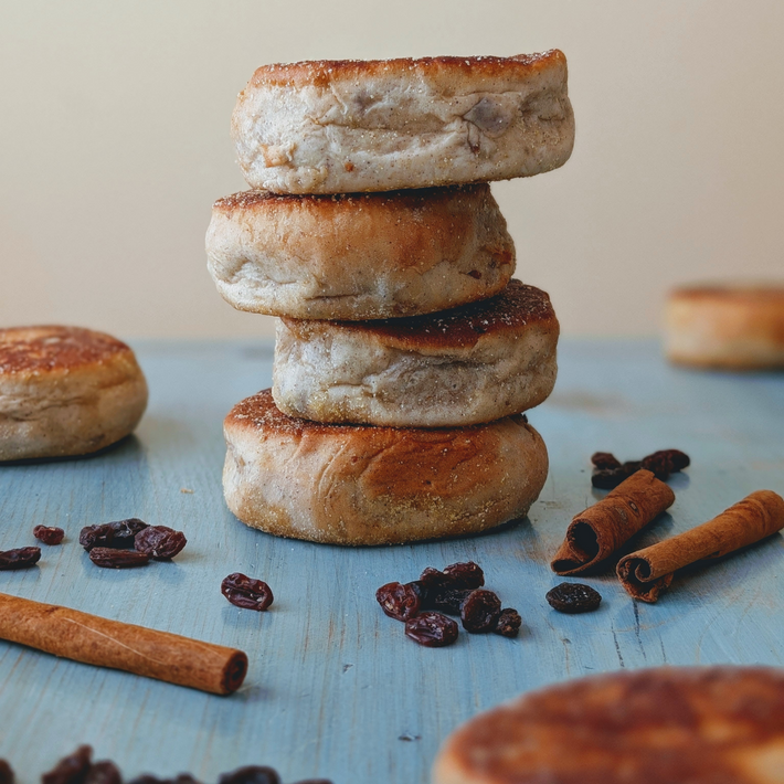 Stack of four toasted English muffins on a blue wooden surface with cinnamon sticks and scattered raisins