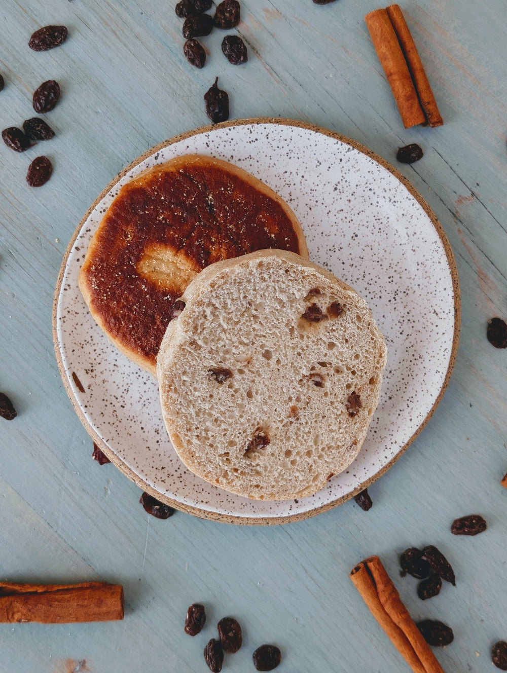 Raisin-studded English muffin halves on a speckled plate, cinnamon sticks and scattered raisins on blue wooden table.