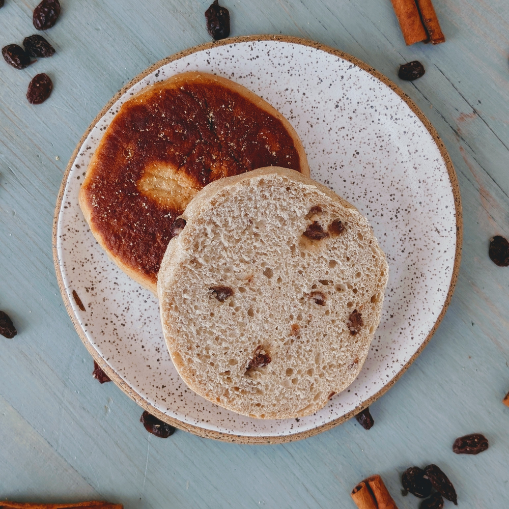 Two raisin buns on a speckled plate, one halved to show raisins; cinnamon sticks and scattered raisins on blue wood.