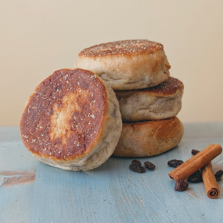 Four toasted English muffins stacked on a blue wooden surface with cinnamon sticks and raisins nearby