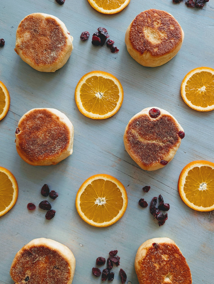 Toasted English muffins and orange slices arranged on blue wood with scattered dried cranberries