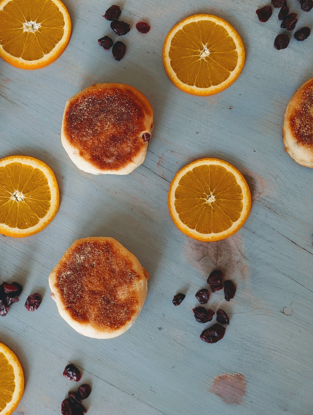 Two toasted English muffins with orange slices and scattered dried cranberries on a pale-blue wooden surface.