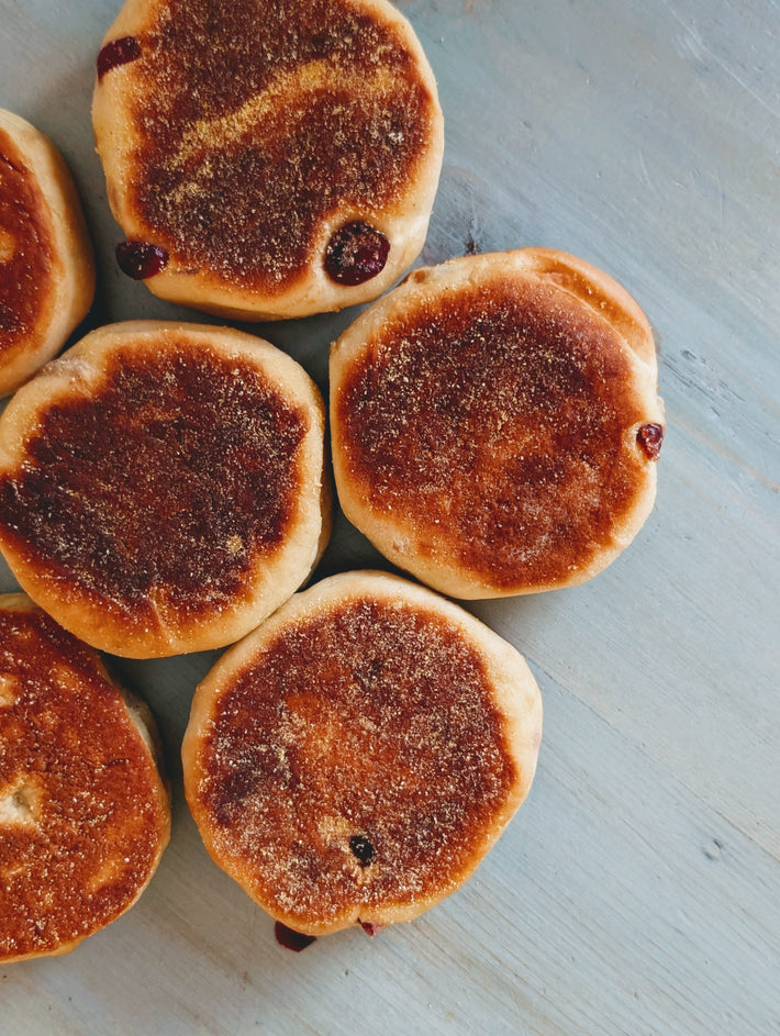 Five toasted English muffins with golden-brown, cornmeal-dusted tops on a pale blue board, some with jam peeking out.
