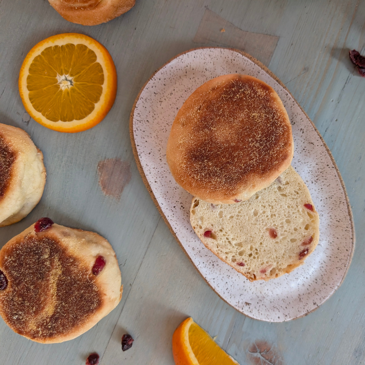 Split cranberry English muffin on speckled oval plate, with whole muffins and orange slices on a light wooden table