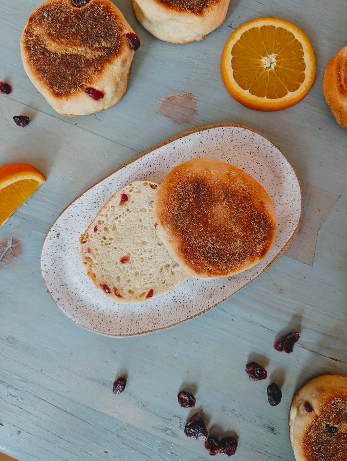 Split cranberry English muffin on speckled oval plate, orange slices and scattered dried cranberries on blue wooden table.