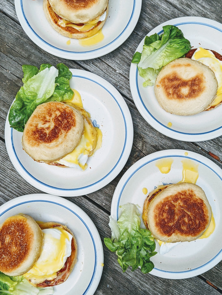 Five English muffin breakfast sandwiches with lettuce, egg, and melted cheese on white plates with blue rims, arranged on a wooden surface.