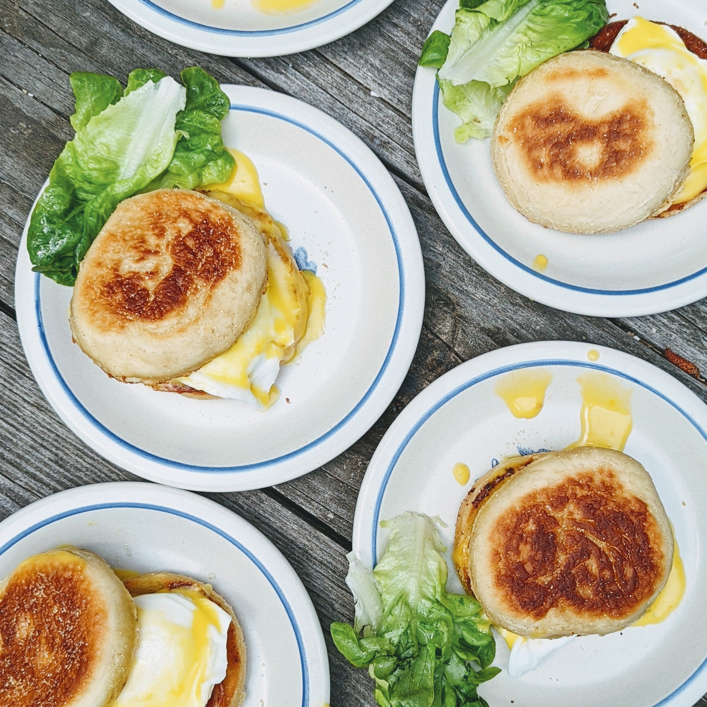Four plates of English-muffin breakfast sandwiches with melted egg, cheese and lettuce on a weathered wooden table.
