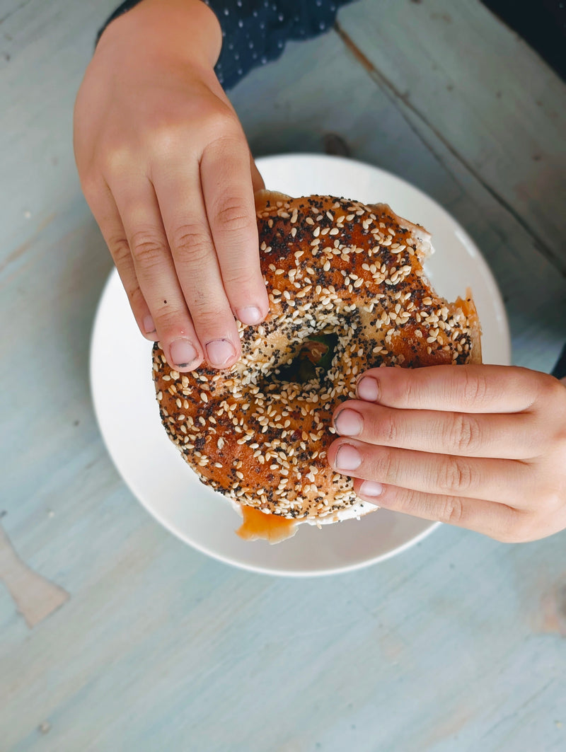 Hands holding an everything bagel topped with sesame and poppy seeds over a white plate on a light blue wooden table