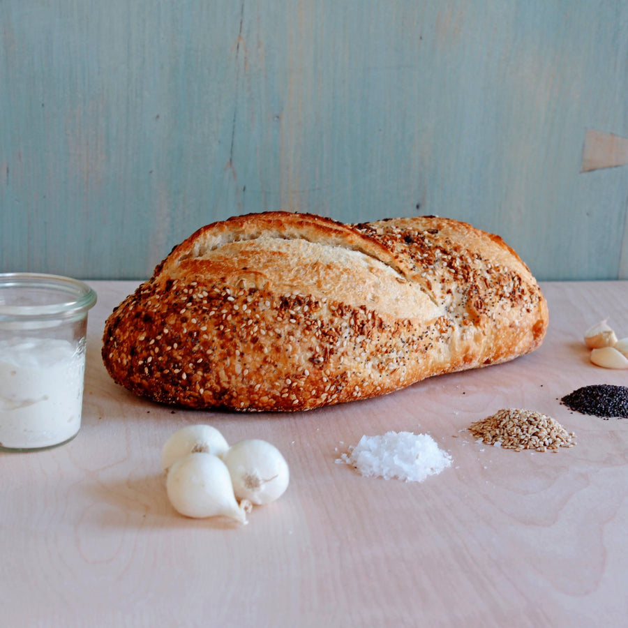 Angled view of Everything Sourdough loaf with seeded crust on a wood surface with garlic, salt, seeds, and flour jar