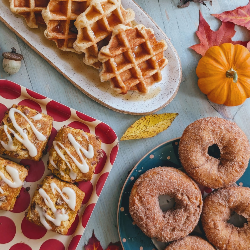 Autumn spread: maple waffles, cinnamon-sugar donuts, and iced pumpkin pastries with fall leaves and a mini pumpkin.