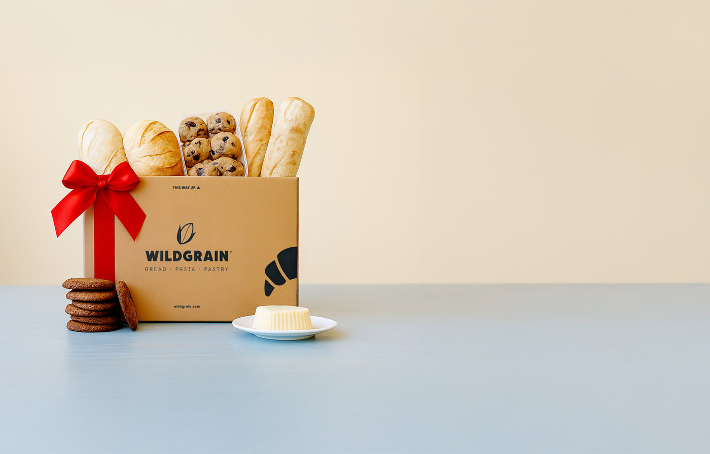 Gift box with a red bow filled with baguettes, chocolate chip cookies, and a plate of butter on a pale backdrop