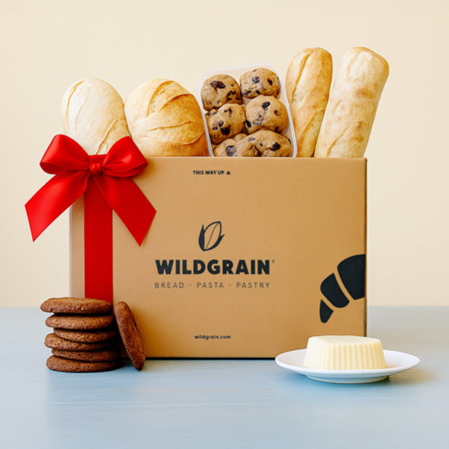Box with bread, cookies, butter and red ribbon; text: 'WILDGRAIN' 'BREAD · PASTA · PASTRY' 'wildgrain.com' 'THIS WAY UP ▲'