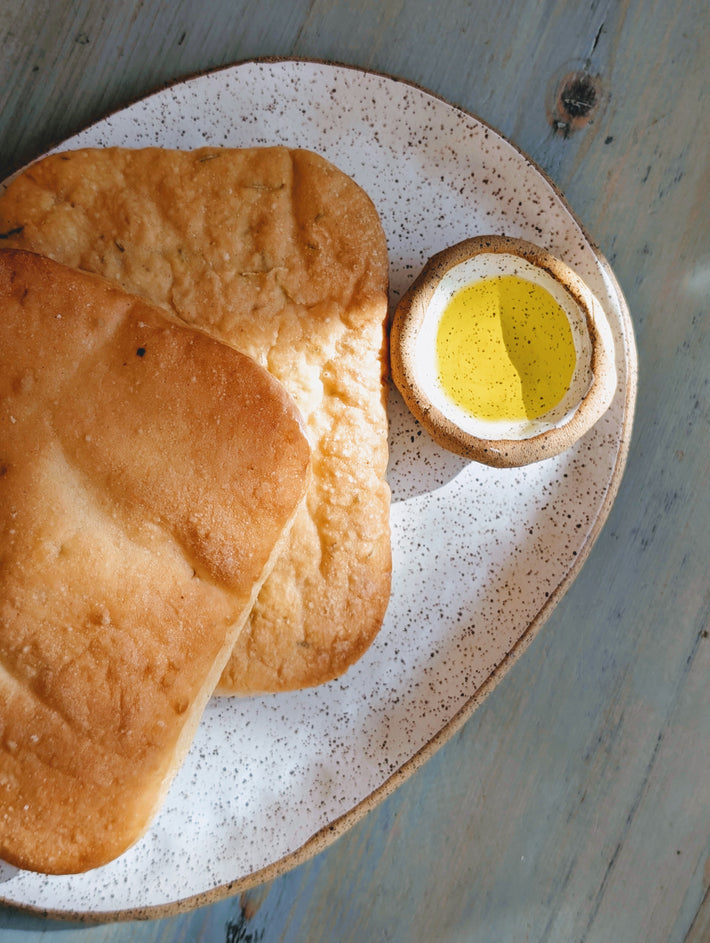 Two toasted flatbreads stacked on a speckled ceramic plate beside a small bowl of olive oil on a light wooden table.
