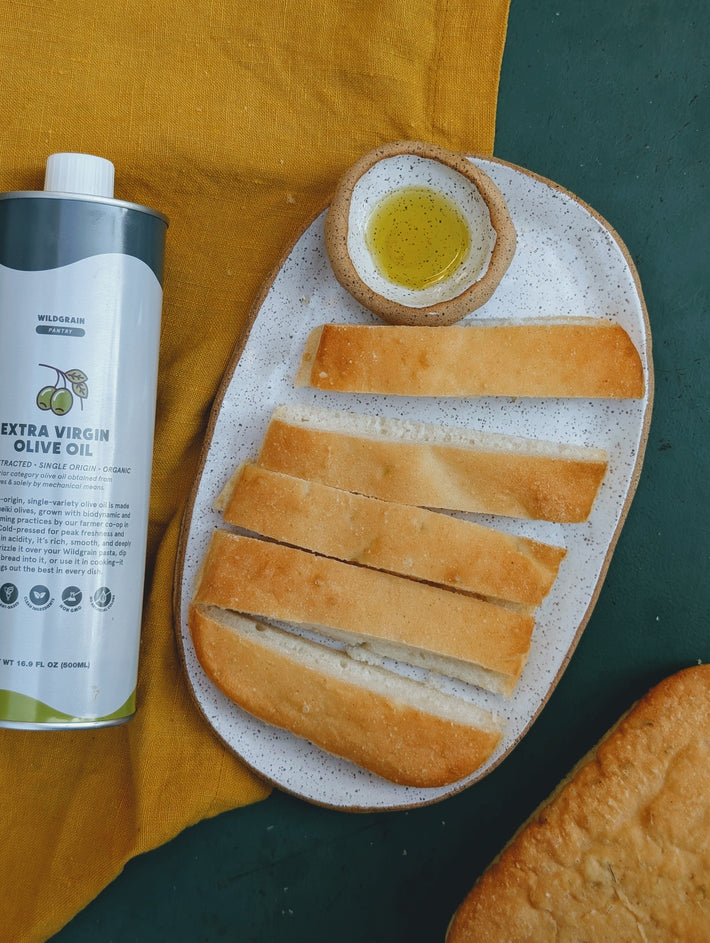 Sliced baguette and small bowl of olive oil on speckled plate beside a can labeled WILDGRAIN PANTRY EXTRA VIRGIN OLIVE OIL