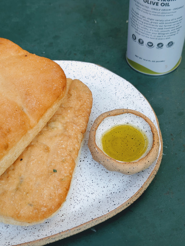 Ciabatta and flatbread on a speckled plate with a small bowl of olive oil; can labeled OLIVE OIL and NET WT 16.9 FL OZ (500ML).