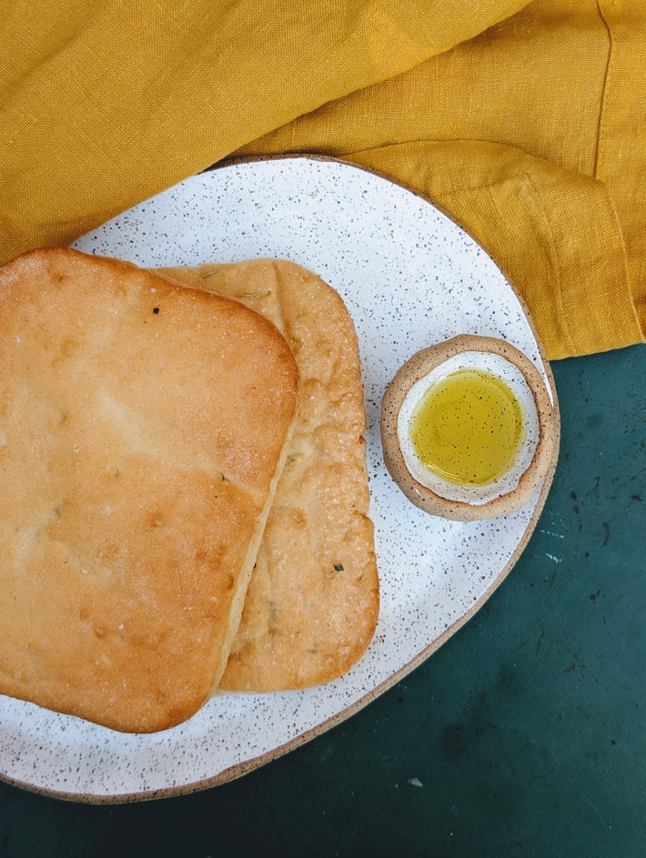 Two square focaccia on a speckled plate with small bowl of olive oil and mustard-yellow napkin.