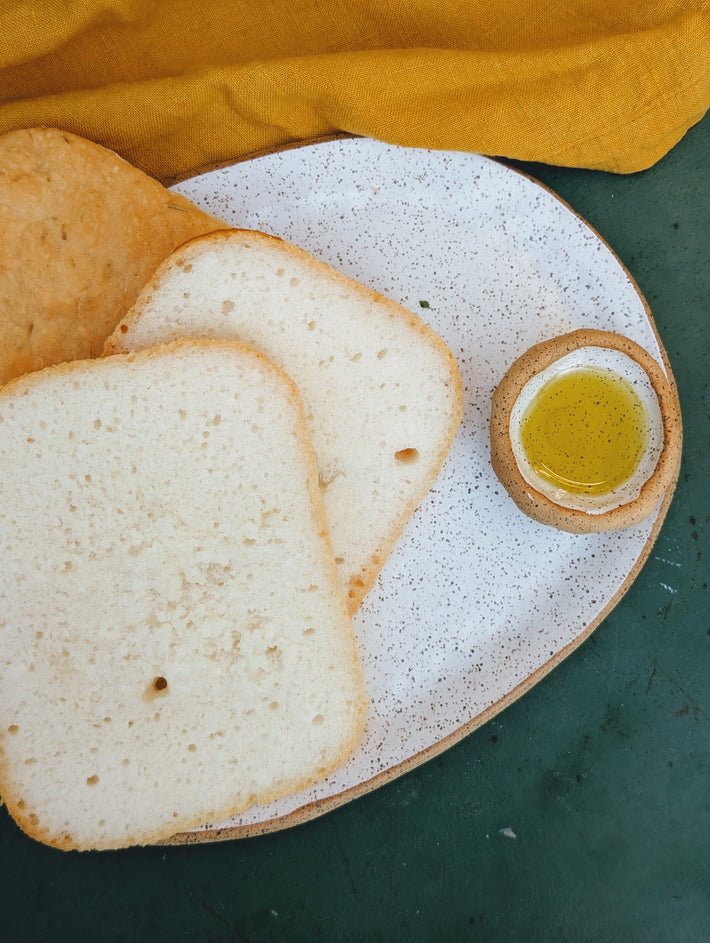 Three slices of white bread on a speckled oval plate with a small bowl of olive oil and a mustard-yellow cloth