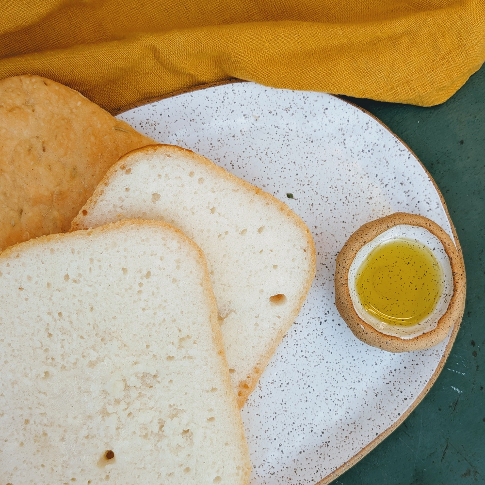 Three slices of gluten-free focaccia on a speckled plate with a small bowl of olive oil and a mustard napkin.