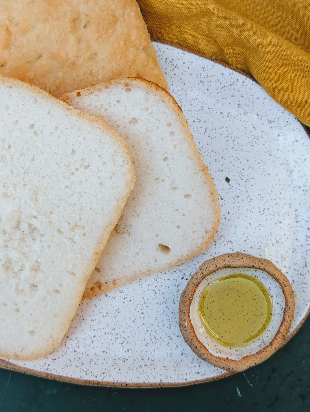 Two slices of white sandwich bread on a speckled ceramic plate with a small dish of olive oil and a mustard-yellow napkin.