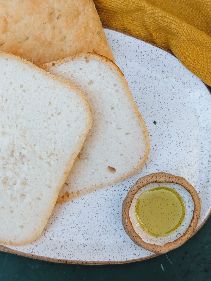 Two slices of white sandwich bread on a speckled ceramic plate with a small dish of olive oil and a mustard-yellow napkin.